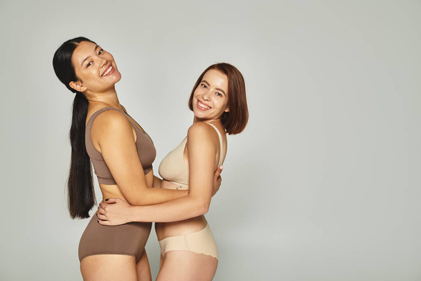 šťastný mladý multikulturní ženy ve spodním prádle objímání navzájem na šedém pozadí, tělo pozitivní - Fotografie, Obrázek