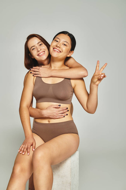 Ευτυχισμένη γυναίκα αγκαλιάζει ασιατικό φίλο της που δείχνει v σημάδι σε γκρι φόντο, το σώμα θετικό - Φωτογραφία, εικόνα