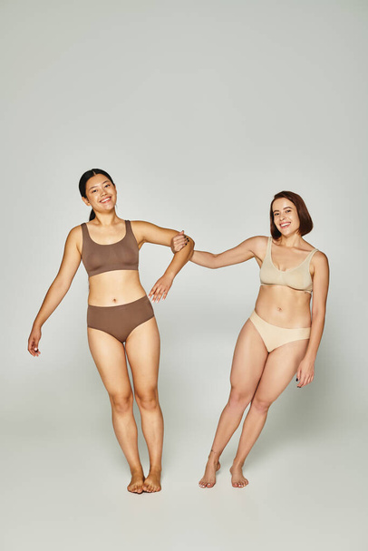 young woman holding hand on asian female friend in underwear while posing together on grey backdrop - Photo, Image