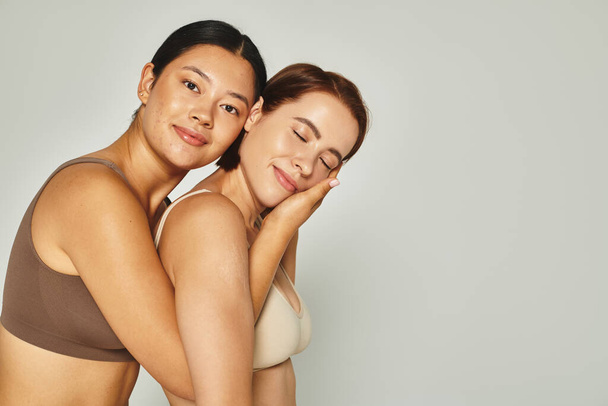 feliz asiático mulher abraçando com ela fêmea amigo em cueca enquanto posando no cinza pano de fundo - Foto, Imagem