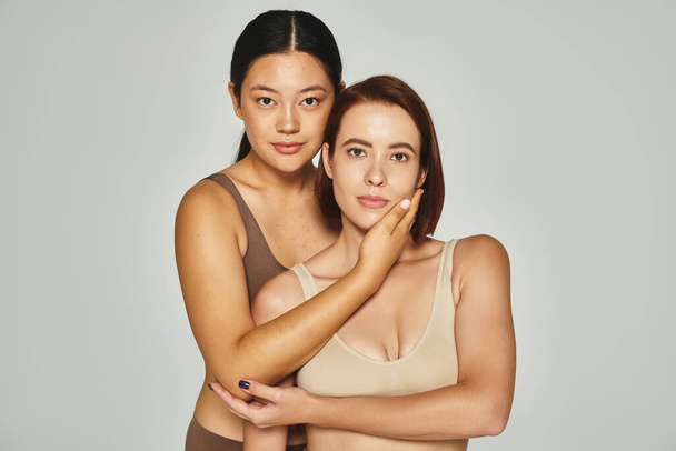 молодая азиатская женщина обнимается со своей подругой в нижнем белье, позируя на сером фоне - Фото, изображение
