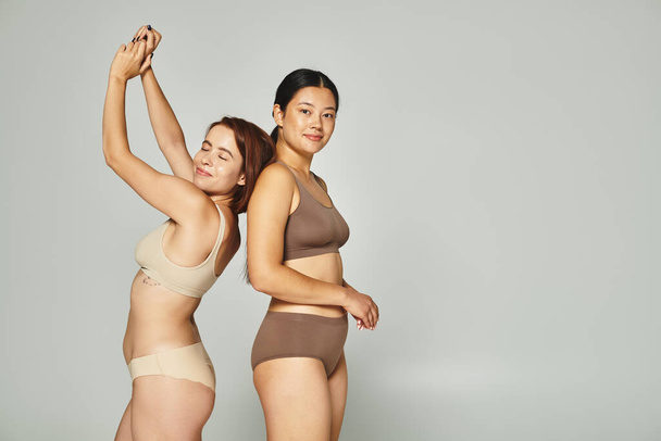 onnellinen nainen alusvaatteet tanssia lähellä aasialainen ystävä alusvaatteet harmaalla pohjalla, kehon positiivinen - Valokuva, kuva