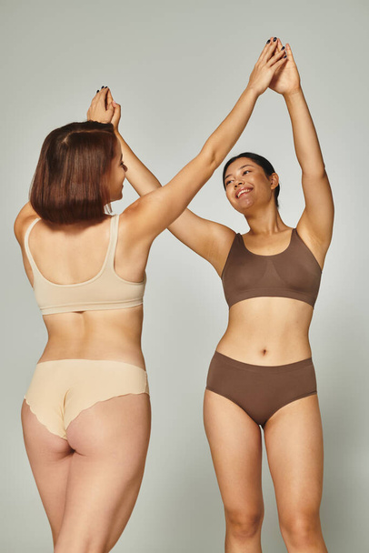 jóvenes feliz interracial mujeres en lencería high-fiving entre sí en gris fondo, cuerpo positivo - Foto, imagen