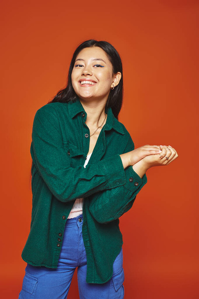 Сияющая молодая азиатская женщина позирует в ярком наряде и смотрит на камеру на оранжевом фоне - Фото, изображение