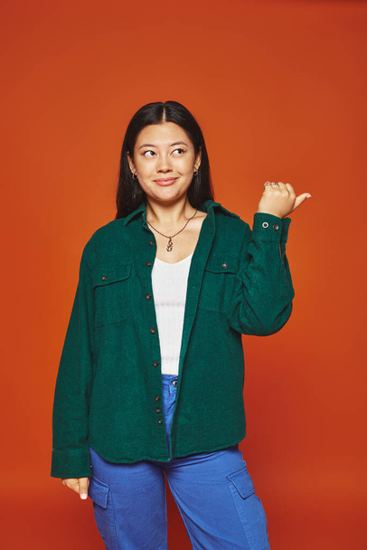 Turuncu arka planda yeşil ceketli canlı poz veren Asyalı kadın. - Fotoğraf, Görsel