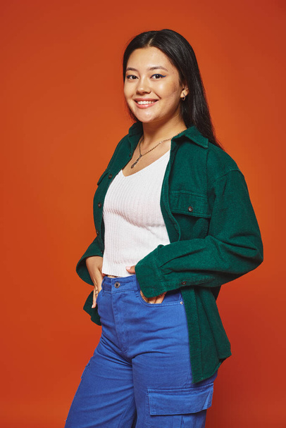 happy young asian woman posing in vibrant outfit with hands in pockets on orange background - Photo, Image
