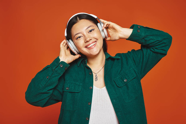 junge und brünette asiatische Frau genießt Musik mit drahtlosen Kopfhörern auf orangefarbenem Hintergrund - Foto, Bild
