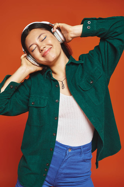 pleased brunette asian woman enjoying music with wireless headphones on orange background - Foto, Imagem