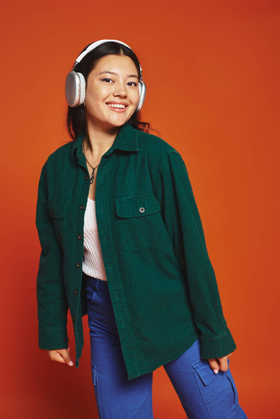 笑顔のブルネット アジアの女性 オレンジ色の背景にワイヤレスヘッドフォンで音楽を楽しんで - 写真・画像