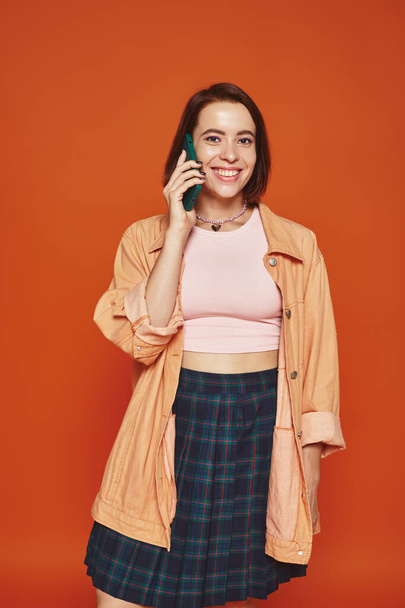 cheerful young woman in vibrant attire talking on smartphone on orange background, phone call - Photo, Image