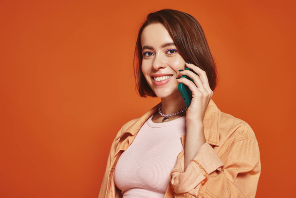 Şık giyinmiş, turuncu arka planda akıllı telefondan konuşan pozitif genç bir kadın. - Fotoğraf, Görsel