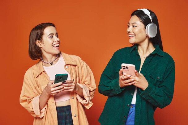 İki mutlu çok kültürlü arkadaş akıllı telefon kullanıyor ve turuncu arka planda müzik dinliyor. - Fotoğraf, Görsel
