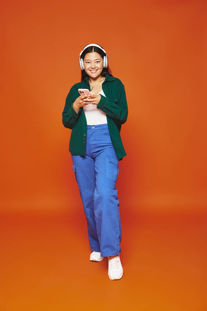 feliz asiático mujer escuchar música en inalámbrico auriculares celebración de teléfono móvil en naranja fondo - Foto, Imagen