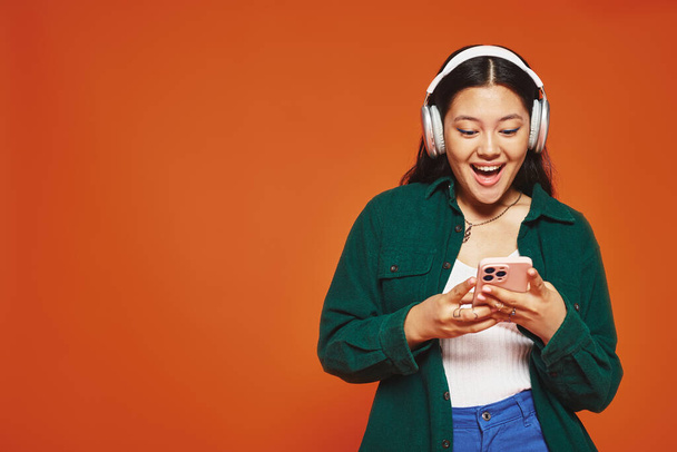 eccitata asiatico donna ascolto musica in wireless cuffie utilizzando smartphone su arancio sfondo - Foto, immagini