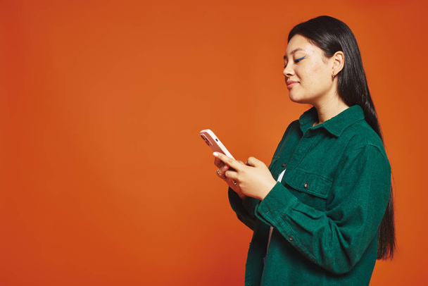 joyful and young asian woman with brunette hair using smartphone and smiling on orange background - Photo, Image
