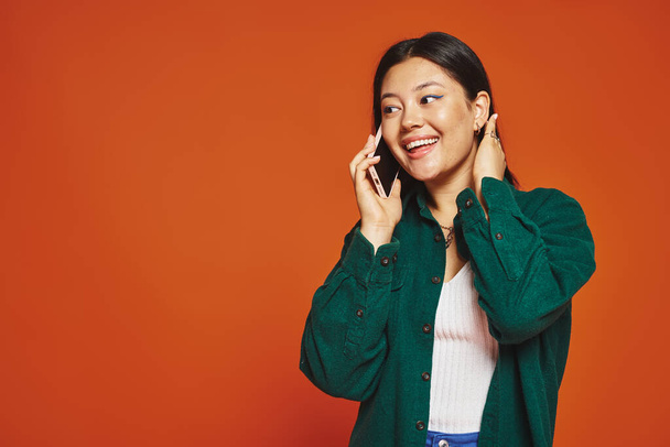 vrolijk jong aziatisch vrouw met brunette haar praten op smartphone en glimlachen op oranje achtergrond - Foto, afbeelding