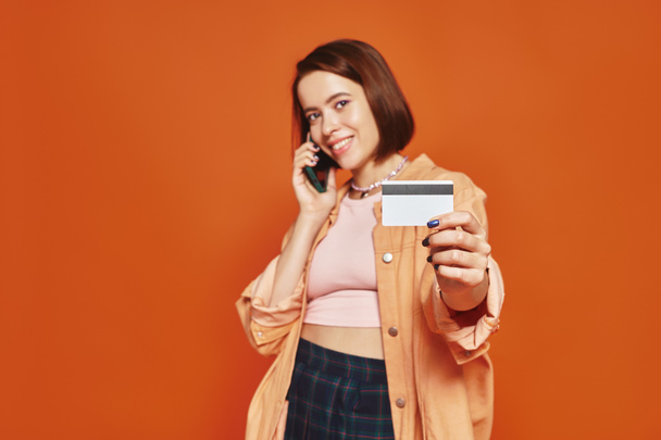 陽気な女性がスマートフォンで話し,オレンジ色の背景にクレジットカードを持っている,オンラインショッピング - 写真・画像
