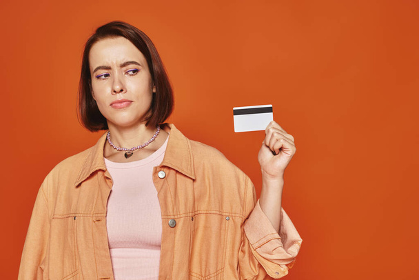 giovane donna premurosa con capelli corti in possesso di carta di credito su sfondo arancione, finanza personale - Foto, immagini