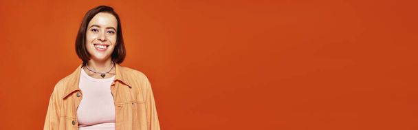 веселая молодая женщина с короткими волосами, смотрящая в камеру и улыбаясь на оранжевом фоне, баннер - Фото, изображение