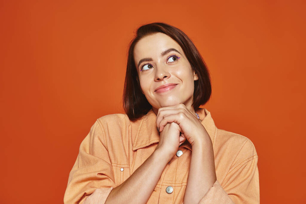 hopeful young woman with short hair smiling and looking away on orange background, cheerful - Photo, Image