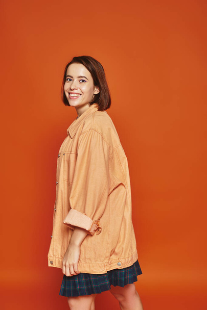 mujer joven positiva con el pelo corto mirando a la cámara y sonriendo sobre fondo naranja, despreocupada - Foto, Imagen