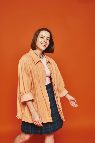 fröhliche junge Frau mit kurzen Haaren, die in die Kamera blickt und auf orangefarbenem Hintergrund lächelt, unbeschwert - Foto, Bild
