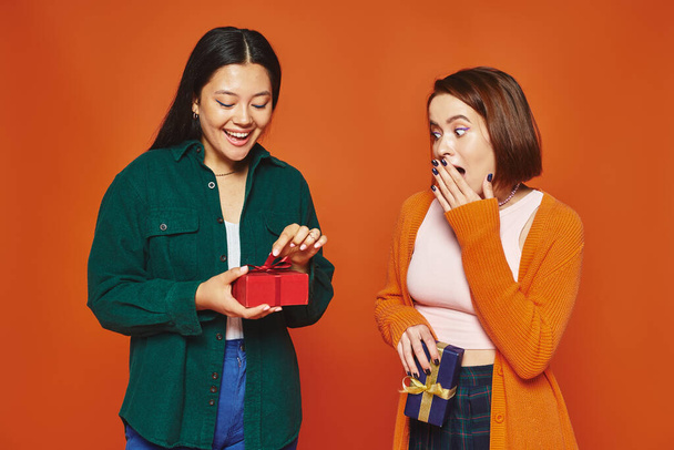 young woman covering mouth and looking at asian friend opening present on orange background - 写真・画像