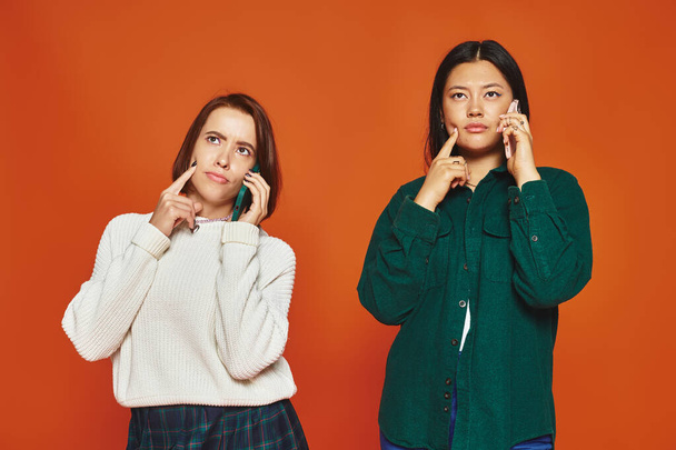 nachdenkliche multikulturelle Frauen in lässiger und lebendiger Kleidung, die auf Smartphone auf orangefarbenem Hintergrund miteinander reden - Foto, Bild