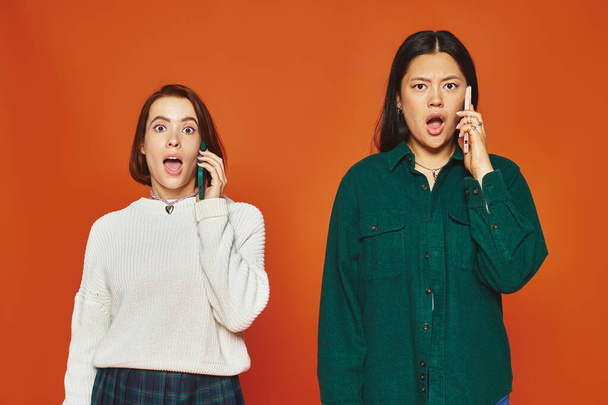 zwei schockierte multikulturelle Frauen in lebendiger Kleidung, die auf orangefarbenem Hintergrund über Mobiltelefone sprechen - Foto, Bild
