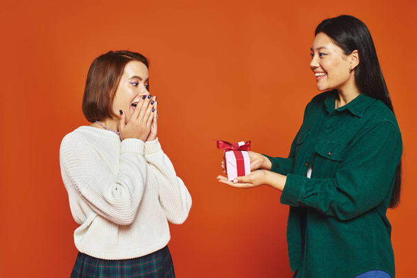 szczęśliwy Azji kobieta dając prezent dla kobieta przyjaciel pokrycie twarz z niespodzianka na pomarańczowym tle - Zdjęcie, obraz