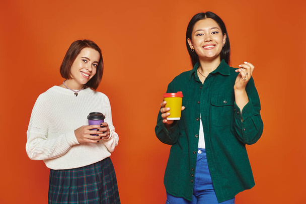 オレンジ色の背景に行くためにコーヒーと紙コップを保持している陽気な多文化女性の友人 - 写真・画像