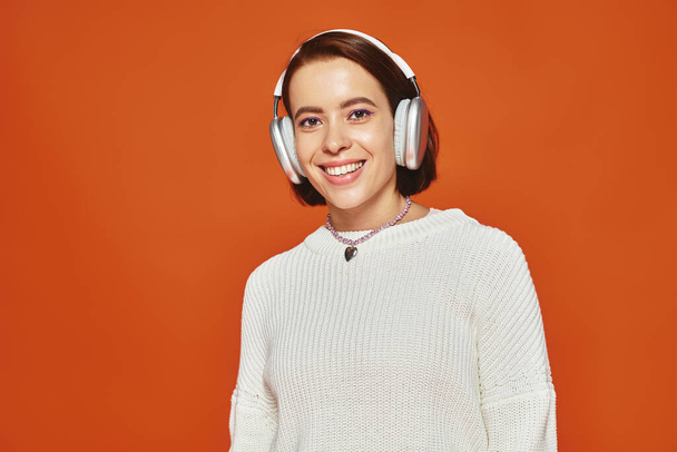 χαρούμενη νεαρή γυναίκα σε λευκό πουλόβερ και ασύρματα ακουστικά ακούγοντας μουσική σε πορτοκαλί φόντο - Φωτογραφία, εικόνα