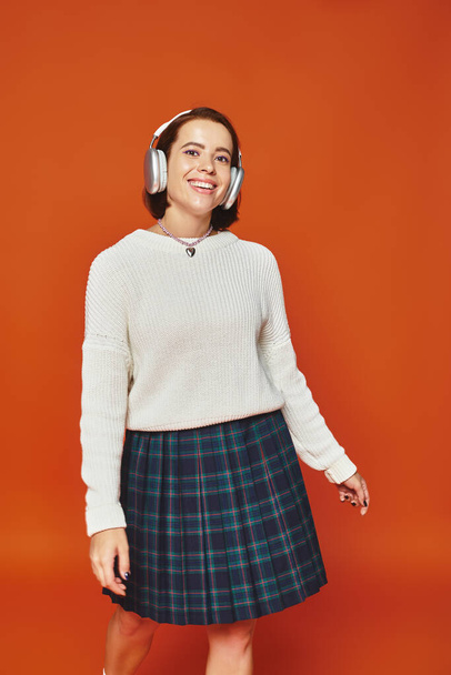 joyful young woman in white sweater and wireless headphones listening music on orange backdrop - Photo, image