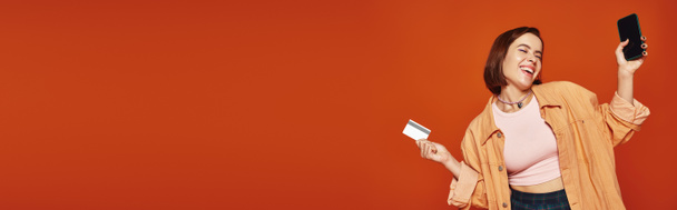 mujer feliz joven sosteniendo teléfono inteligente y tarjeta de crédito sobre fondo naranja, banner de compras en línea - Foto, imagen