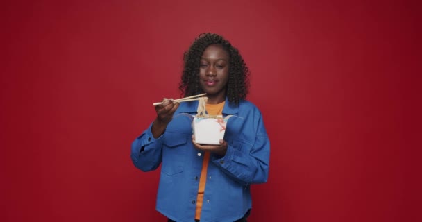 Afro-Amerikaanse vrouw kijkt naar camera en glimlacht, bereidt zich voor om noedels, zeevruchten en groenten te eten met eetstokjes op geïsoleerde rode achtergrond. Aziatisch straatvoedsel. Vaste fastfood klaar voor levering. - Video