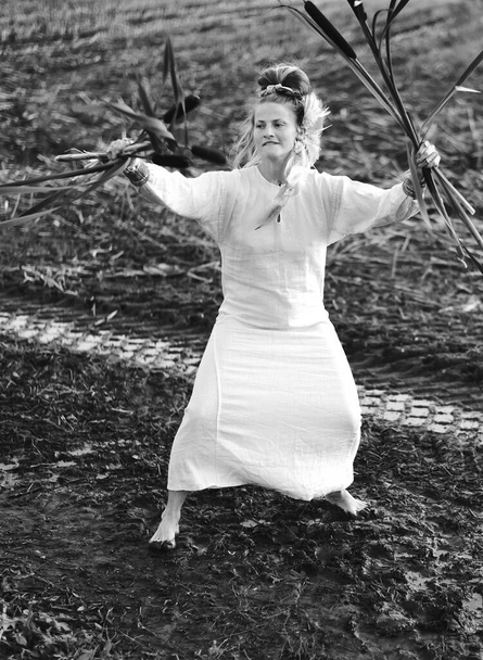 Красива жінка в сукні танцює босоніж на брудному полі, тримаючи очерет в руках - Фото, зображення