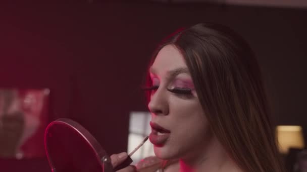 Medium lähikuva valkoihoinen queer mies kirkas meikki, väärä ripset ja peruukki soveltamalla huulikiilto valmistautuessaan vetää show backstage huoneessa neon punainen valo - Materiaali, video
