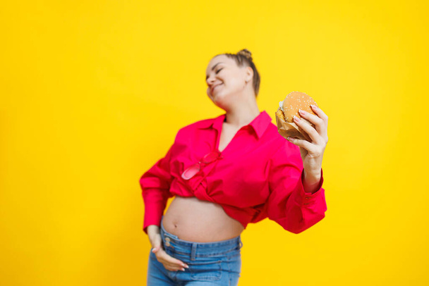 Μια χαριτωμένη έγκυος με ροζ πουκάμισο τρώει ένα φαστφουντάδικο. Μια έγκυος γυναίκα σε κίτρινο φόντο με ένα ορεκτικό μπέργκερ στα χέρια της. Επιβλαβής τροφή για έγκυες γυναίκες. - Φωτογραφία, εικόνα