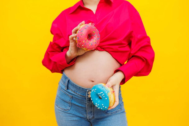 黄色の背景にピンクのシャツを着た明るい妊婦が甘いドーナツを食べている. 妊娠した女性の手の新鮮な甘いドーナツ - 写真・画像