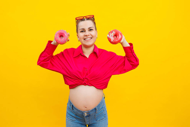 Яскрава вагітна жінка в рожевій сорочці на жовтому тлі їсть цукерки. Солодкі пончики в руках вагітної жінки. - Фото, зображення