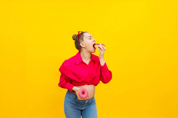 A terhes nő édes fánkot eszik sárga alapon. Egy terhes nő rózsaszín ingben és szemüvegben édes tortát eszik. Káros étel a terhesség alatt. - Fotó, kép