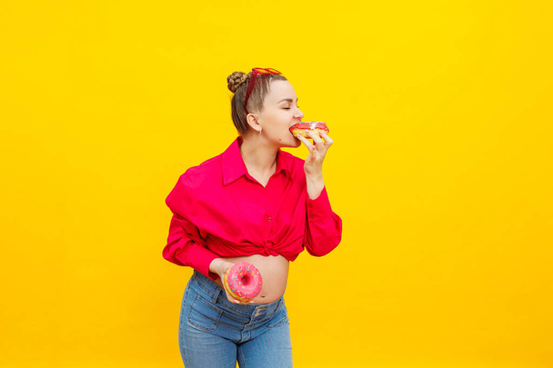 mujer regente come rosquillas dulces sobre un fondo amarillo. Una mujer embarazada con una camisa rosa y gafas se come un pastel dulce. Alimento nocivo durante el embarazo. - Foto, imagen