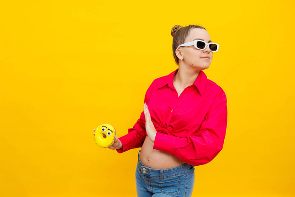 Zwangere vrouw eet zoete donuts op een gele achtergrond. Een zwangere vrouw in een roze shirt en bril eet een zoete taart. Schadelijk voedsel tijdens de zwangerschap. - Foto, afbeelding