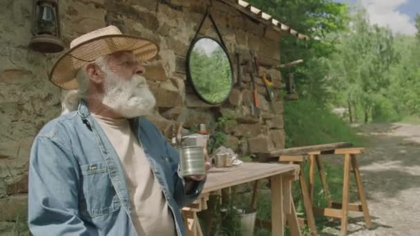 Plan moyen de pensionné masculin détendu dans un chapeau buvant du café assis à l'extérieur d'une petite maison en brique - Séquence, vidéo