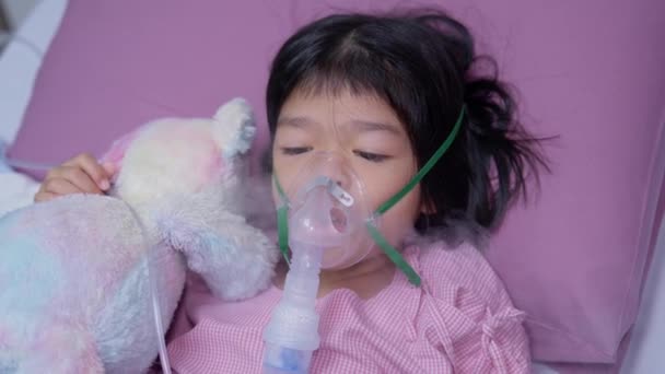 Egy ázsiai kislánynak oxigénmaszkja van, és porlasztón keresztül lélegzik a kórházban. Fogalma hörghurut, légzőszervi és orvosi kezelés, inhalációs orvoslás, köd, egy porlasztó. - Felvétel, videó