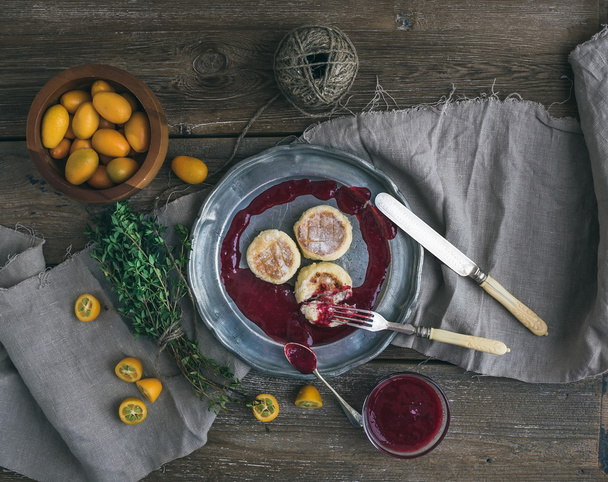 Rustic breakfast set. Russian cheese cakes on a vintage metal plate with lingonberry jam, fresh kumquats, thyme, decoration rope and old dinnerware over rough wooden desk - Photo, image