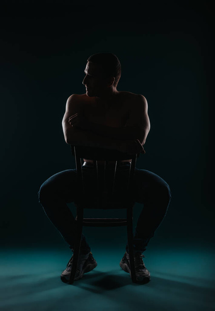Strong macho looking aside, sitting on a chair. Male fitness instructor posing in studio. Silhouette photo - Photo, Image