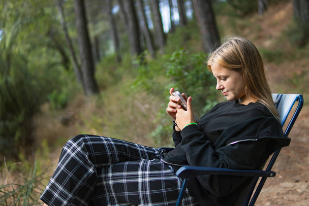 a teenage girl in casual clothes sits on a chair in a Camping and looks at the phone, a girl sits in the forest among the trees on a folding chair. High quality photo - Фото, зображення