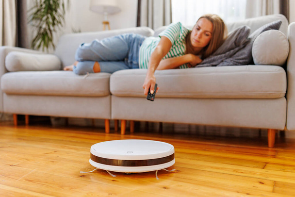 Robot aspiradora limpiar una habitación mientras una mujer se relaja en el sofá. Una mujer opera un vacío con un mando a distancia - Foto, Imagen