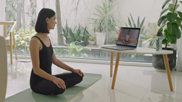 Жінка сидить на тренувальному килимку і розмовляє з інструктором фітнесу через онлайн-відеодзвінок на ноутбук, маючи особисте тренування вдома - Кадри, відео